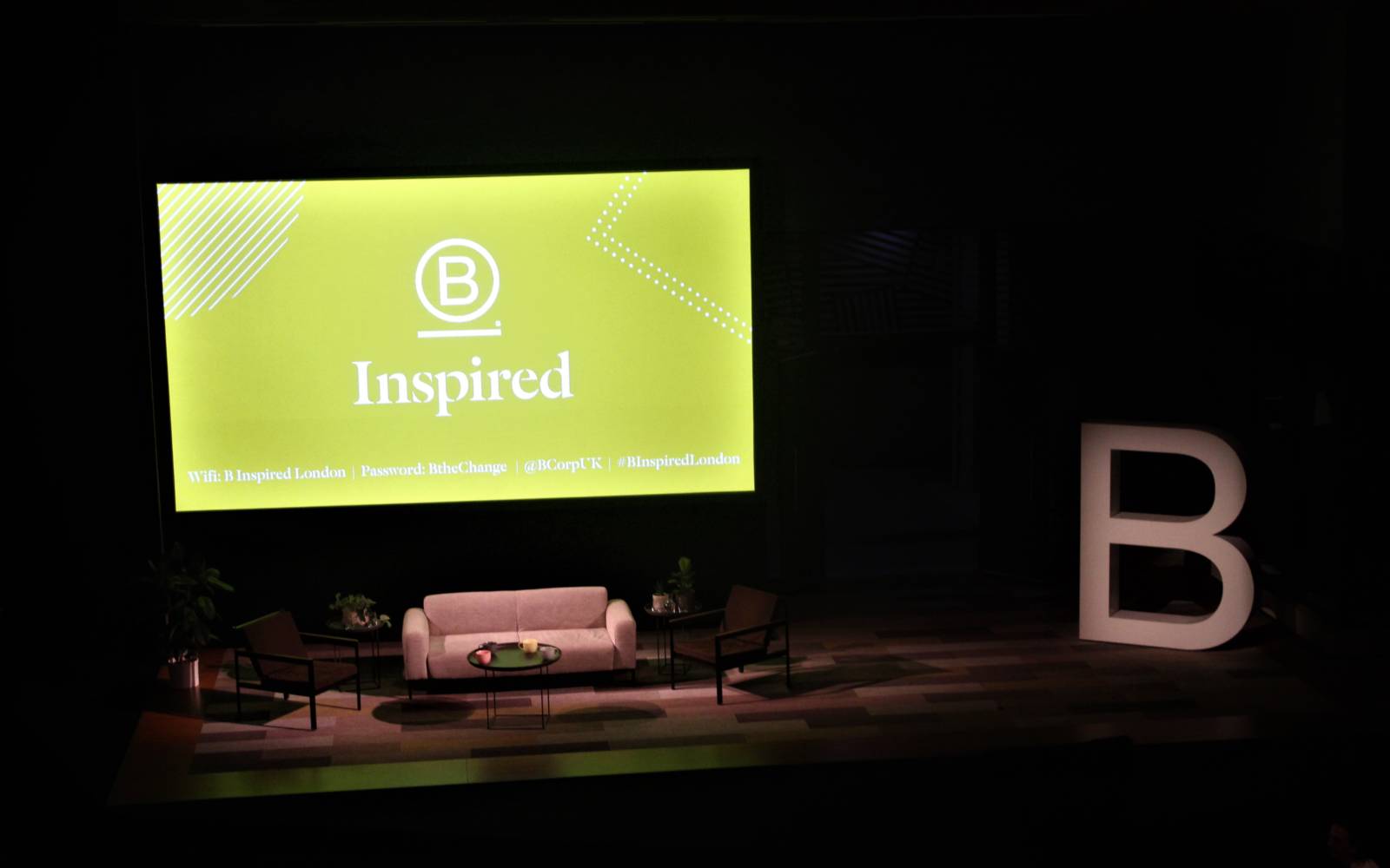 Photo of Bridge Theatre Stage. There is a big screen at the bag that has a green background and reads B Inspired. There is a sofa in front of this and a large B standing next to it.