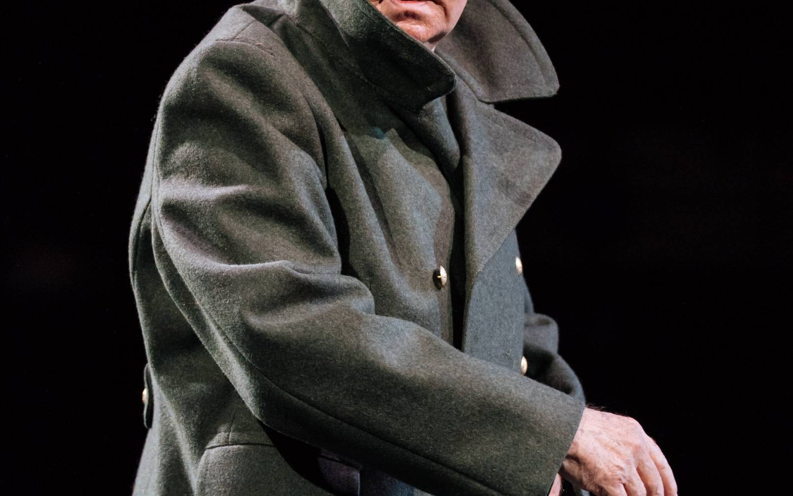 An old man, wearing a coat with the collar turned up, looks behind him, concerned