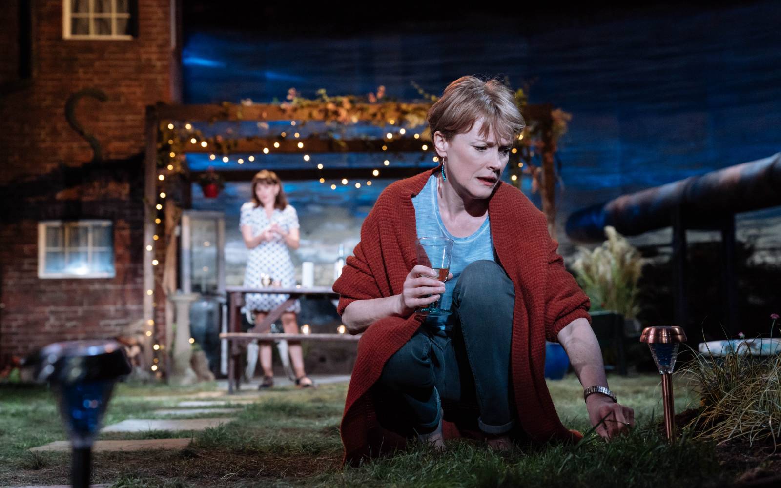 Night. A woman crouches, wine glass in hand, inspecting a patch of grass. In the background, a younger woman looks on