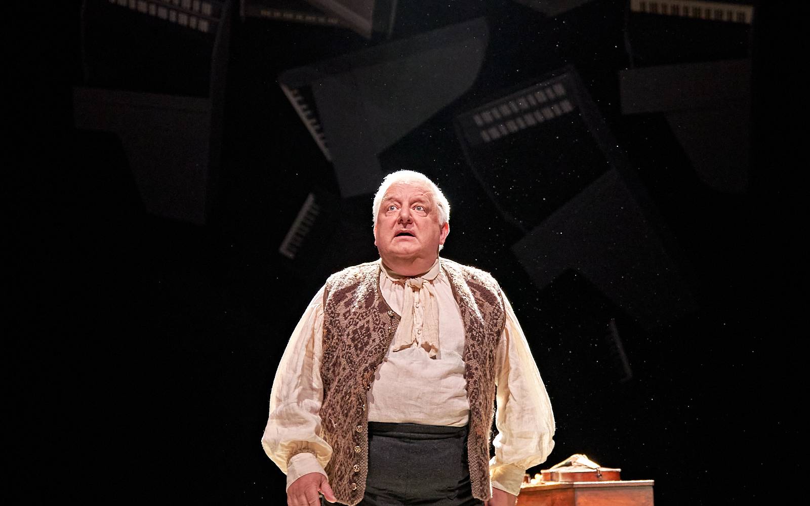 Simon Russell Beale, with short grey hair and dressed in 18th-century clothing, stands in front of a wooden keyboard instrument. Above his head, black harpsichords are suspended from the ceiling.