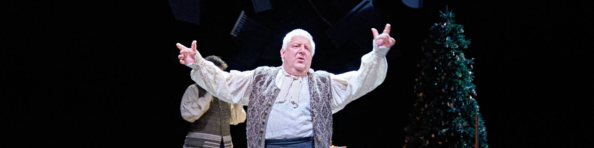 Simon Russell Beale, with short, grey hair and wearing 18th-century dress, dances in front of a wooden keyboard instrument. In the background, a Christmas tree.