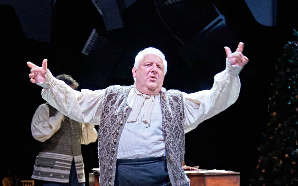Simon Russell Beale, with short, grey hair and wearing 18th-century dress, dances in front of a wooden keyboard instrument. In the background, a Christmas tree.