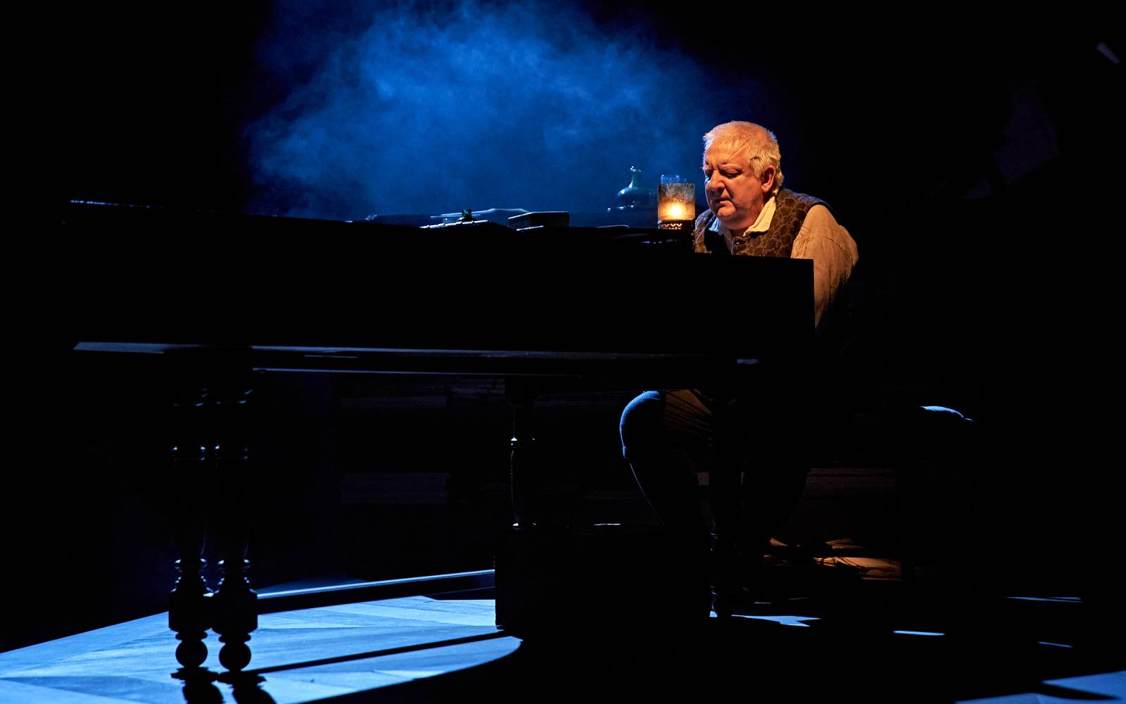 On a dark stage, lit by candlelight, Simon Russell Beale plays a keyboard instrument.