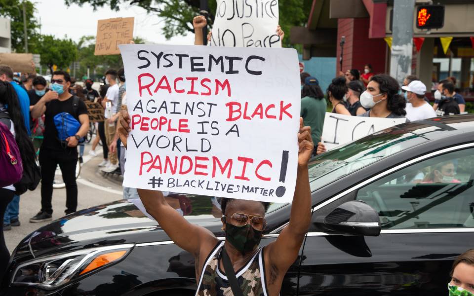 A black man wearing a mask holds up a placard reading 'Systemic racism against black people is a world pandemic! #blacklivesmatter' In the background are other protestors