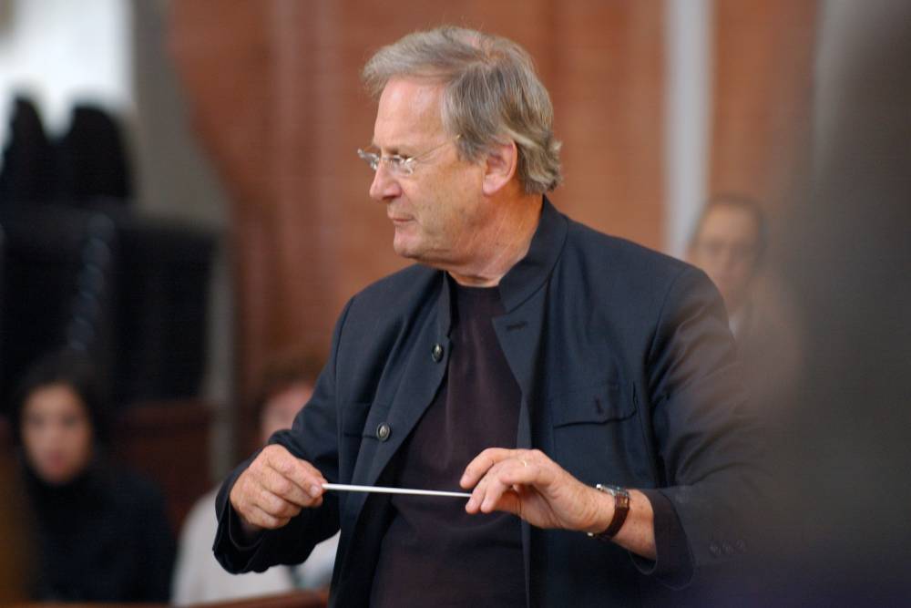 A man with grey hair and glasses, wearing a black shirt and dark blue jacket, holding a conductor's baton.